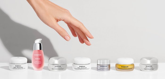 Cinq produits de soins de la peau sur une surface blanche comprennent un sérum rose et quatre pots de crème de formes et couleurs variées, y compris en argent et en blanc.