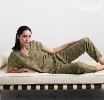 Frau lehnt auf einem cremefarbenen Sofa, trägt grünes florales Oberteil und Hose, mit "Triumph"-Logo in der oberen rechten Ecke.