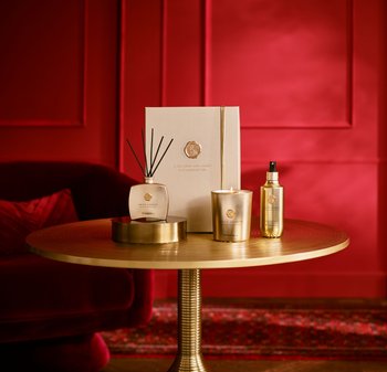 Kobberfargede lys og diffusor, begge på et rundt gyllent bord. Bakgrunnen har en rik rød vegg, med en dekorativ gaveeske i nærheten.