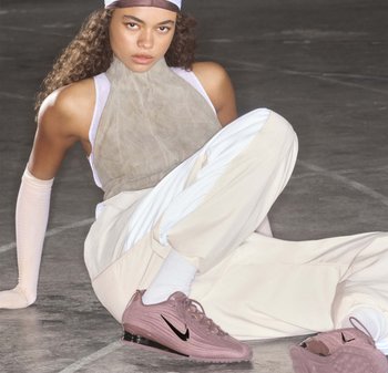 Young woman in beige and white outfit sitting on the floor wearing pink Nike sneakers with black swoosh logo.