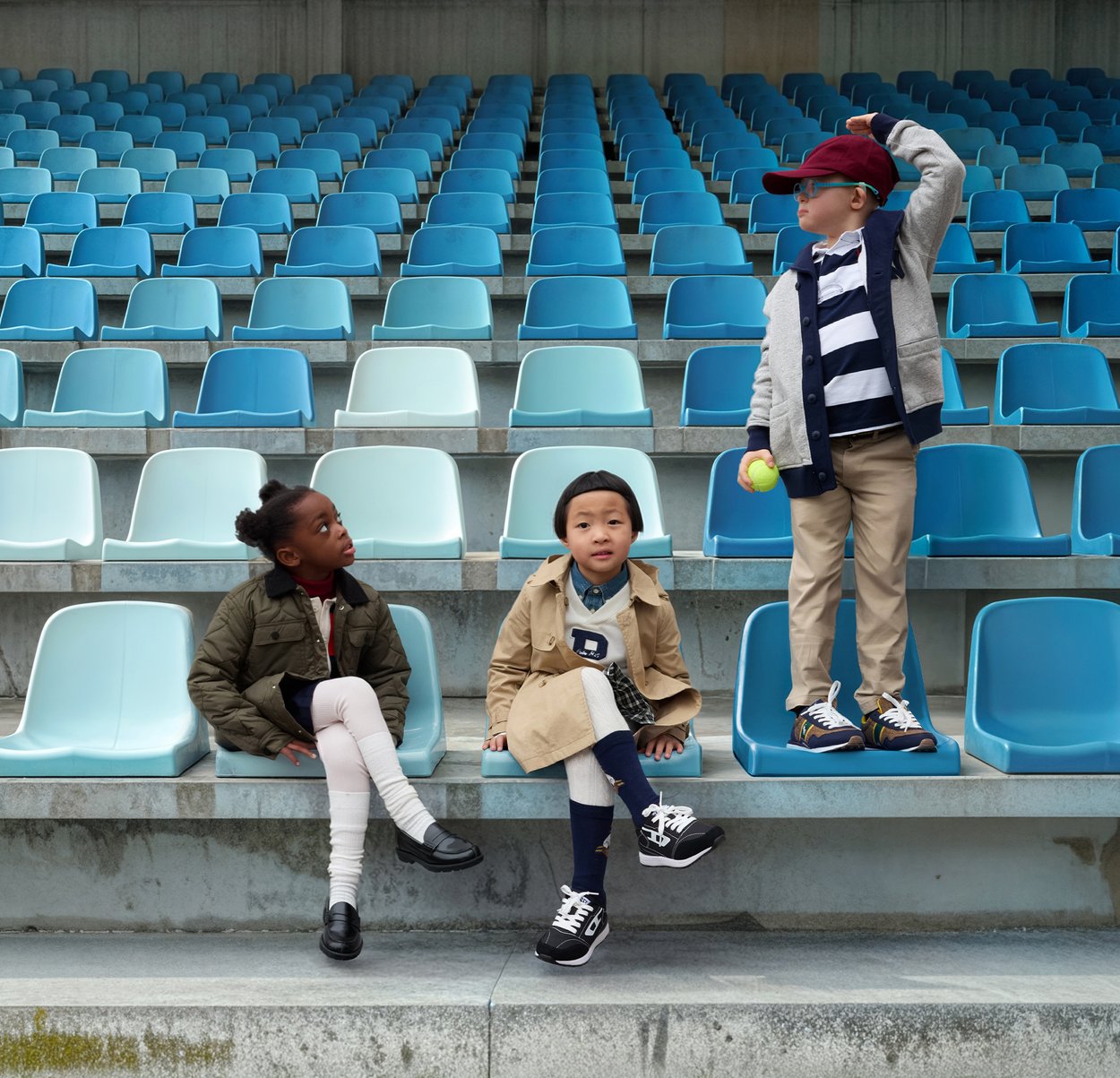 Kolme lasta istuu stadionilla, jossa on sinisiä ja turkooseja penkkejä. Yksi heistä käyttää beigeä trenssitakkia, toinen tummansinistä paitaa ja kolmas punaista lippalakkia.
