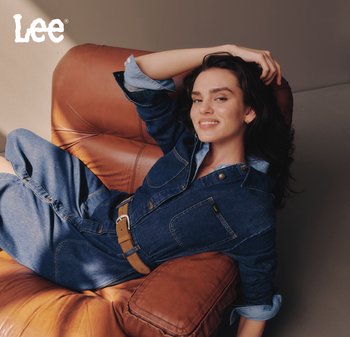 Combinaison en denim avec un col bleu clair et des accents de boutons dorés, associée à une ceinture en cuir brun, assise sur un fauteuil en cuir brun.