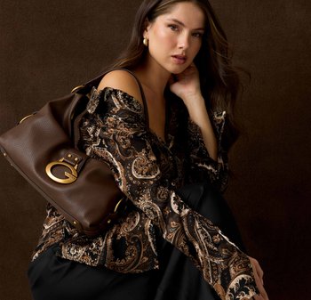 Bruine leren handtas met gouden hardware, gecombineerd met een blouse in zwart en bruin met paisleypatroon. Model zit tegen een donkerbruine achtergrond.
