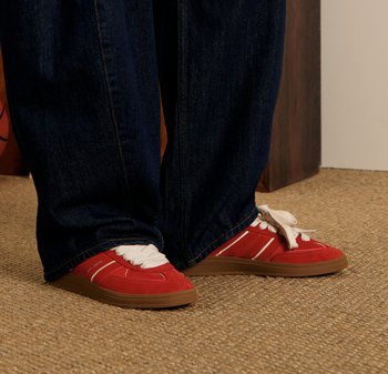 Czerwone zamszowe sneakersy z białymi paskami i sznurówkami, brązowe gumowe podeszwy. Połączone z ciemnoniebieskimi spodniami o szerokich nogawkach, na tle teksturowanego beżowego tła.