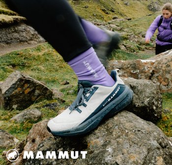 Hellgraue Wanderschuhe mit blauen Akzenten, ausgestattet mit einer strukturierten Sohle, Schnürsenkeln und Logo. Getragen mit lila Socken auf steinigem Terrain.
