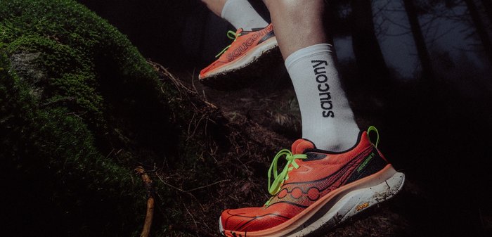 Running shoes in bright orange with black accents and reflective detailing, featuring a cushioned sole and green laces. Socks are white with black logo.