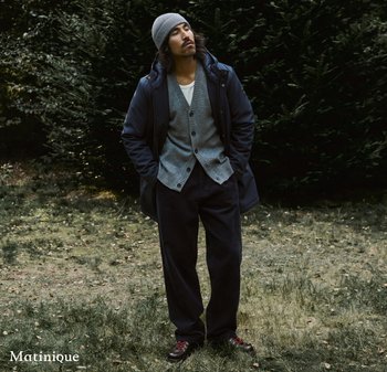 Man in gray beanie, navy jacket, gray cardigan, black pants, and brown hiking shoes stands with hands in pockets on grassy area near trees.