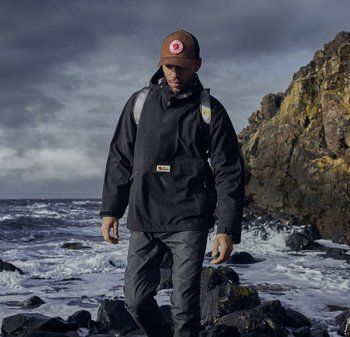 Schwarze wasserdichte Jacke mit einer Fronttasche, die über dunklen Hosen getragen wird. Graue Rucksackriemen und eine braune Kappe mit einem Logo. Felsige Küstenlandschaft im Hintergrund.
