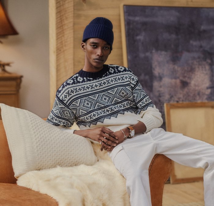Knitted jumper with blue and white geometric patterns, crew neck design. Paired with white trousers, seated on a tan armchair with cushions.