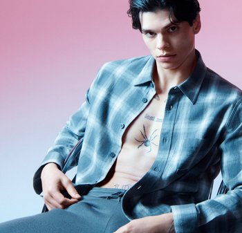 Jasnoniebieska koszula w kratę, rozpięta pod szyją, odsłaniająca ciało z tatuażem w postaci pająka. Gładka tekstura, swobodny krój, na różowym gradientowym tle.