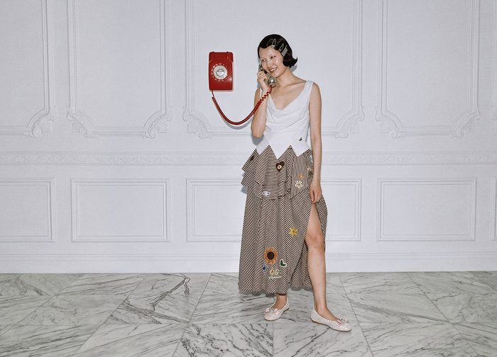 Γυναίκα με κοντά μαλλιά και κλιπς στα μαλλιά μιλάει σε κόκκινο τηλεφωνικό ακουστικό vintage σε τοίχο, φοράει λευκή μπλούζα και φούστα με σχέδιο και σκίσιμο στο πόδι, στέκεται σε μαρμάρινο πάτωμα.