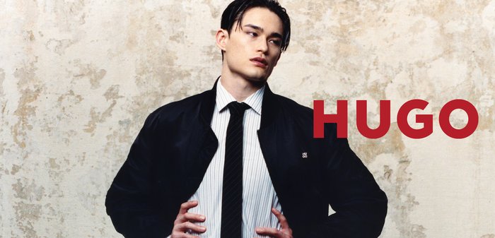 Joven con chaqueta negra, camisa blanca y corbata negra, de pie frente a una pared texturizada con la palabra "HUGO" en letras rojas y negritas.