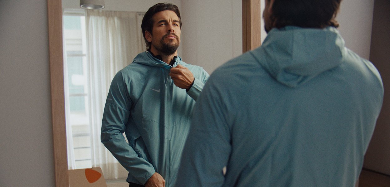 Hombre con una chaqueta azul cerrándola mientras se mira en un espejo grande en una habitación con luz tenue.
