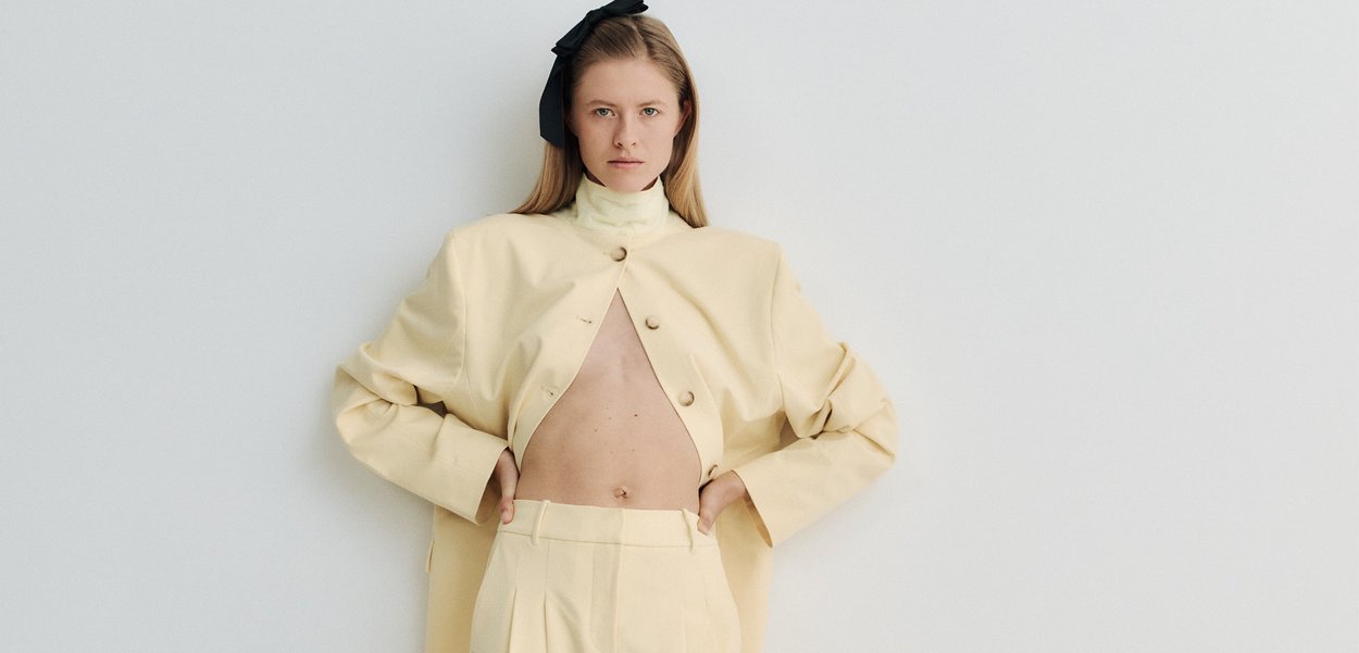 Nainen, jolla on päällään beige ylipitkä takki avoinna paljastaen paljaan vatsan, ja siihen sopivat korkeavyötäröiset housut, seisomassa yksinkertaisen valkoisen taustan edessä.