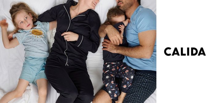 Famiglia di quattro persone sdraiata su un letto, genitori sorridenti, padre che abbraccia il figlio più piccolo, figlio maggiore disteso rilassato in pigiama.