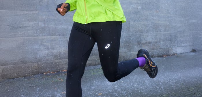 Green lightweight jacket, black leggings with reflective logo, black and orange trainers, purple ankle socks. Wet pavement background.