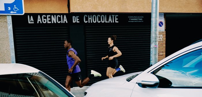 Light beige athletic running shoes with thick, ventilated soles placed on gray asphalt in sunlight and shadow.