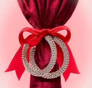 Zilverkleurige, gestructureerde creolen die hangen aan een rode satijnen strik, vastgemaakt rond een bordeauxrode fluwelen stof op een roze achtergrond.