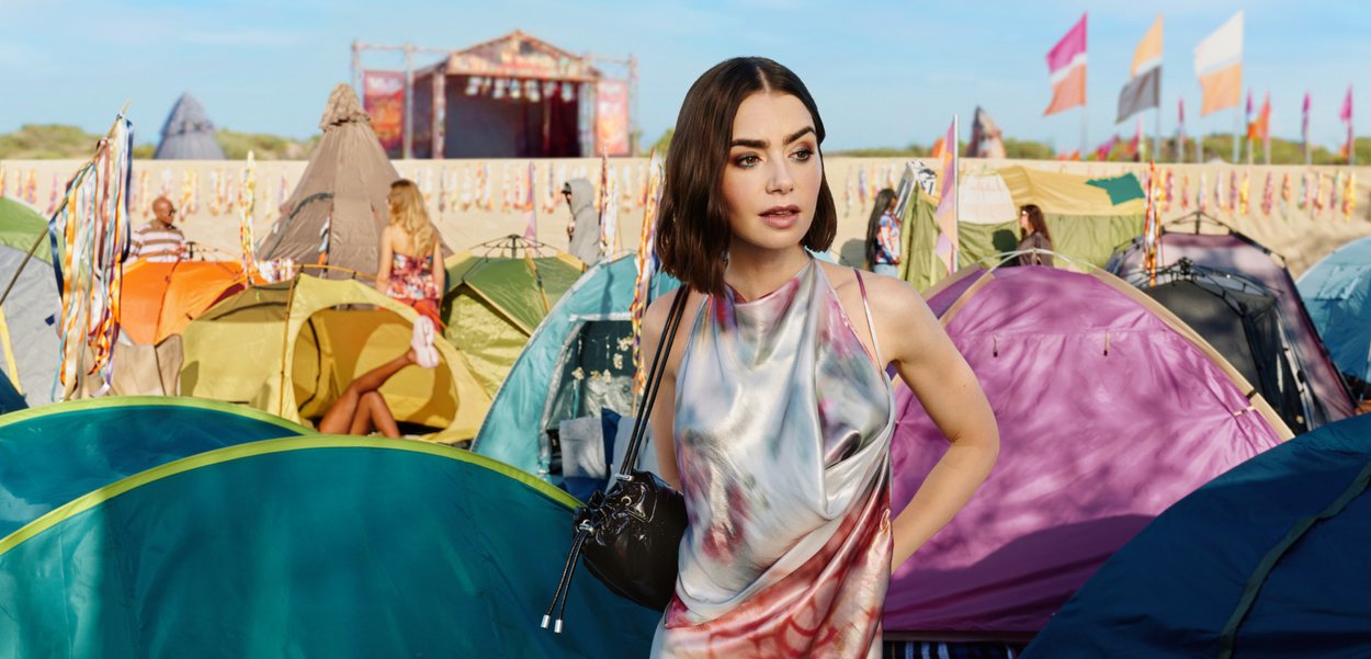 Νεαρή γυναίκα με πολύχρωμο φόρεμα στέκεται ανάμεσα σε ζωηρά χρωματισμένες σκηνές κάμπινγκ σε ένα ηλιόλουστο υπαίθριο φεστιβάλ με σκηνή και σημαίες στο φόντο.