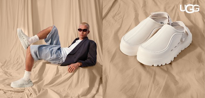 Mann iført hvite slip-on sko, hvite sokker, teksturerte blå shorts, hvit skjorte, svart blazer og solbriller, hvilende på beige stoffoverflate.