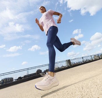 Een persoon die buiten jogt op een zonnige dag, gekleed in roze hardloopschoenen, marineblauwe leggings en een lichtroze T-shirt, bij een metalen reling.