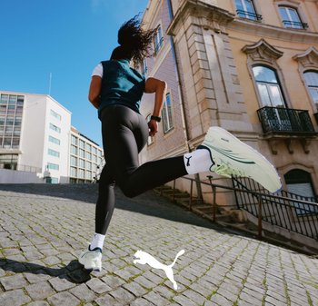 Scarpe da corsa Puma di colore verde chiaro con dettagli bianchi e suole nere. Chi le indossa ha dei leggings neri e sta correndo su una strada acciottolata.