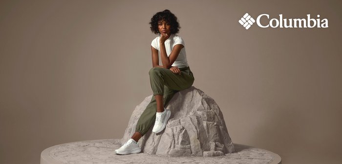Femme en baskets blanches, t-shirt blanc et pantalon vert assise pensivement sur un rocher avec le logo Columbia en arrière-plan.