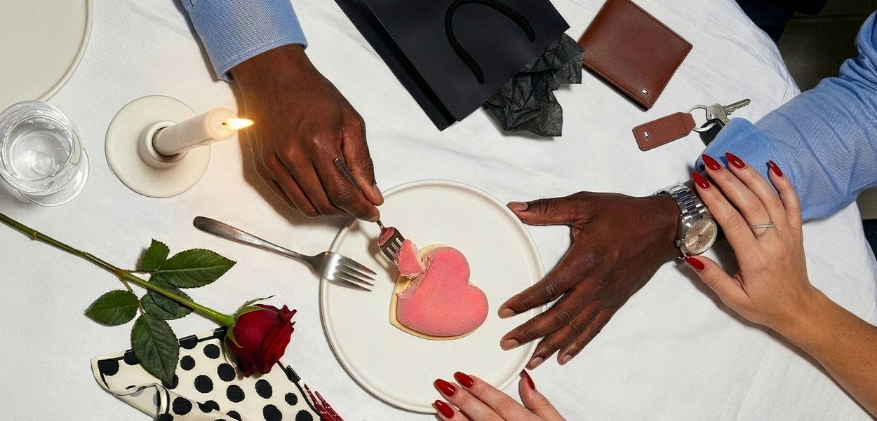 Paar jagab küünlavalgel laual südamekujulist magustoitu, millel on roos, võtmed, rahakott ja sõrmed õrnalt kokku puudutamas.