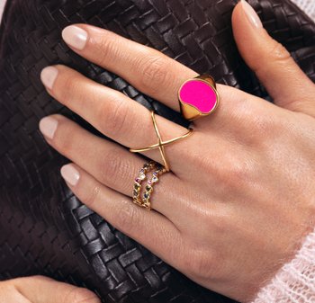 Bagues en or avec un accent rose, un design torsadé, et une bande de pierres multicolores sur une main, sur un fond sombre en tissu tissé.