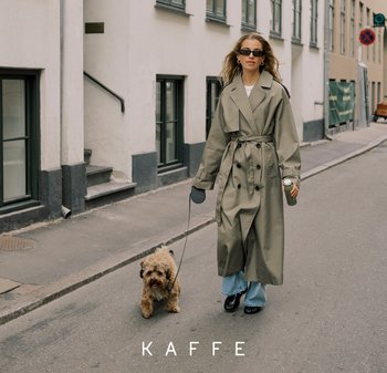 Woman in long green trench coat and sunglasses walking a small dog on leash down a city street, holding a drink cup in one hand.