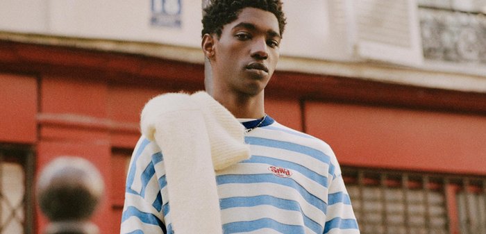 Jeune homme portant un pull rayé bleu et blanc avec une écharpe blanche sur l'épaule, debout dans un environnement urbain.