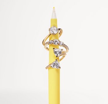 Vier goldene Ringe, die auf einem gelben Bleistift gestapelt sind, mit verschiedenen Diamantformen: rund, herzförmig und dreieckig, und unterschiedlichen Texturen der Ringe.