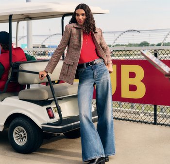 Gepunkteter Blazer in Beige und Rot, rotes Polohemd, weit geschnittene blaue Jeans, schwarzer Gürtel und Plateaustiefel, posiert neben einem Golfwagen.