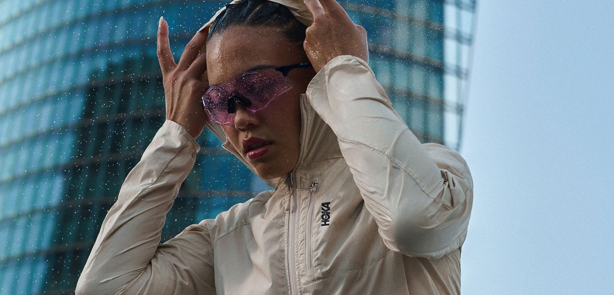 Personne portant une veste beige clair et des lunettes de sport violettes ajuste la capuche avec un bâtiment en verre en arrière-plan.