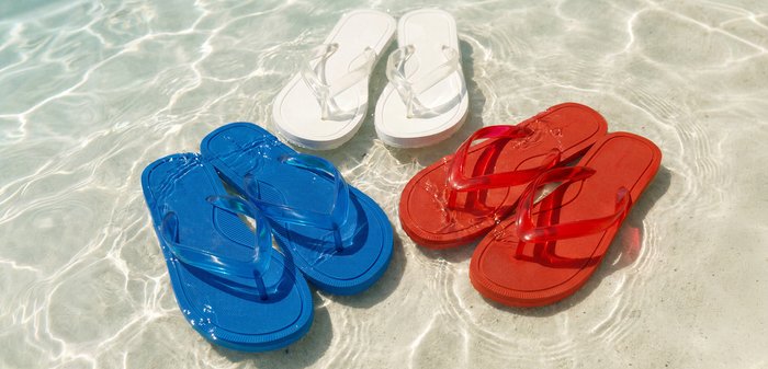 Three pairs of flip-flops in blue, white, and red with clear straps resting in shallow, clear water with sunlight reflections.