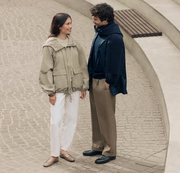 Giacca a cappuccio beige con vita elastica, abbinata a pantaloni bianchi. L'uomo indossa una sciarpa blu scuro e pantaloni, con scarpe marroni. Sfondo in pavimentazione di pietra.