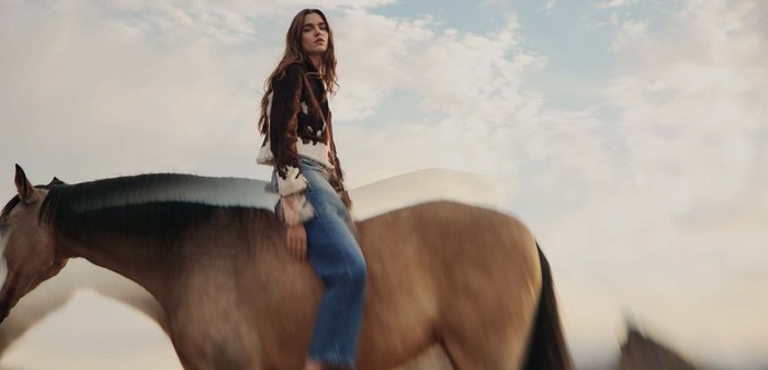 Femme aux cheveux longs portant une veste à motifs et un jean, assise en amazone sur un cheval brun sous un ciel partiellement nuageux.