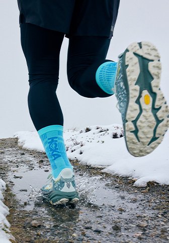 Αθλητικά παπούτσια τρεξίματος με γκρι και μπλε διχτυωτό επάνω μέρος, υφή στη σόλα και ορατό πέλμα, συνδυασμένα με μπλε κάλτσες και μαύρα κολάν, σε μια βρεγμένη, χιονισμένη διαδρομή.