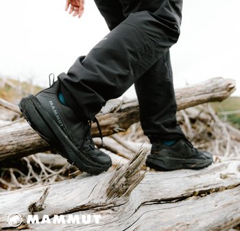 Schwarze Outdoor-Schuhe mit strukturiertem Sohle und verstärkten Bereichen, kombiniert mit dunklen Hosen, auf einem Stamm in einer natürlichen Umgebung platziert.