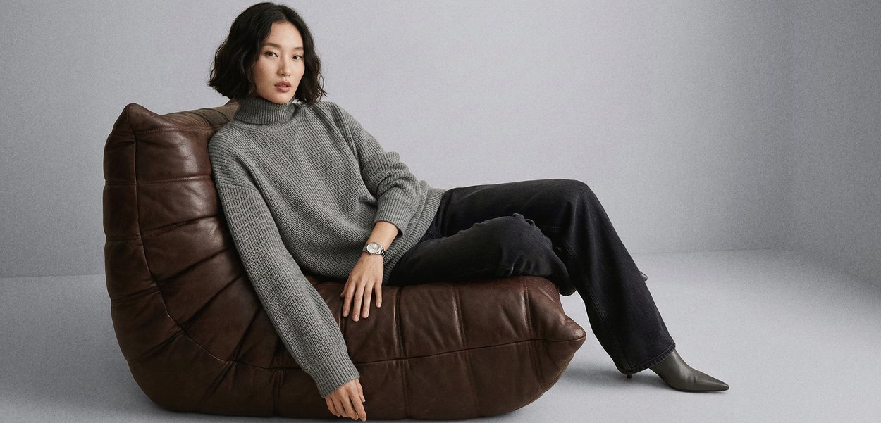 Woman with black hair reclining on a brown leather cushion chair, wearing a grey turtleneck jumper, black trousers, grey boots, and a silver watch.