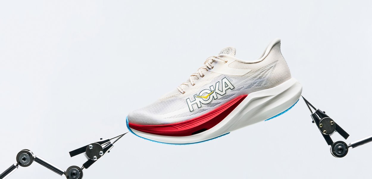Chaussure de course HOKA beige clair et rouge tenue par des bras mécaniques sur un fond blanc uni.