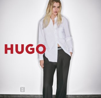 Weißes Hemd mit Knopfleiste und kleiner Tasche sowie dezentem Logo, kombiniert mit dunklen, weit geschnittenen Hosen. Der Hintergrund ist schlicht weiß.