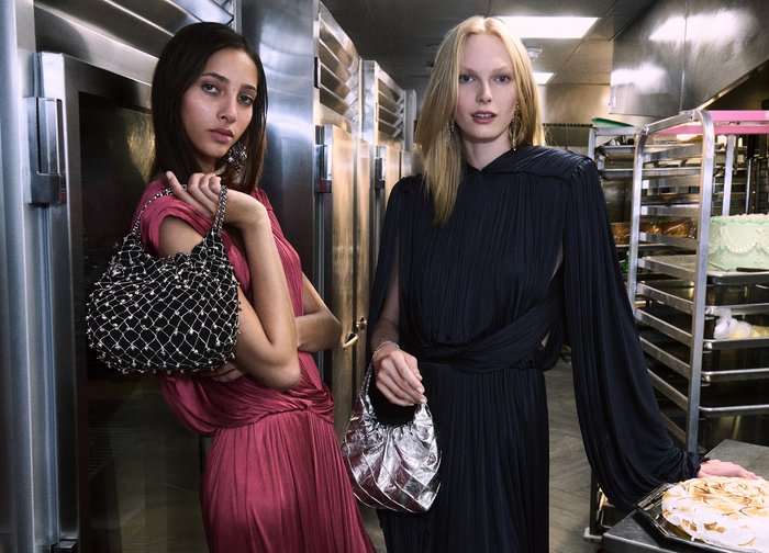 Två modeller i ett kök: en bär en svart och silverprydd väska; den andra en glänsande silverväska. Båda klänningarna är flödande, i rosa och mörkblått.