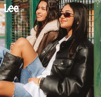 Dos mujeres están sentadas una al lado de la otra contra una valla de metal verde, una lleva una chaqueta de borrego, la otra viste una chaqueta de cuero negra y gafas de sol.
