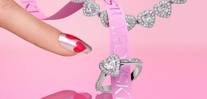 Hartvormige diamanten ketting en ring op een roze achtergrond met een roze lint en een hand met nagelkunst in hartthema.