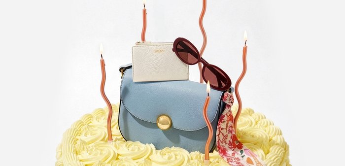 Μπλε δερμάτινη τσάντα με χρυσό κούμπωμα, λευκό πορτοφόλι, κόκκινα γυαλιά ηλίου και κασκόλ πάνω σε κίτρινη τούρτα με παγωμένη επιφάνεια και πέντε αναμμένα στριφογυριστά κεριά.