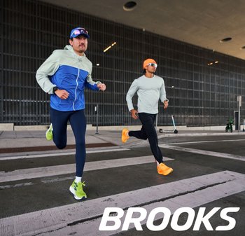 Due corridori uomini in abbigliamento sportivo corrono su una strisce pedonali. Uno indossa una giacca azzurra e blu scuro, l'altro una maglia grigia e pantaloni neri. Scarpe luminose.