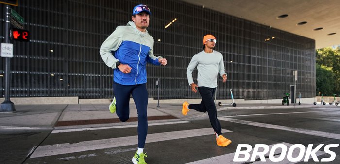 Due corridori sono in movimento su una strisce pedonali. Uno indossa una giacca blu e verde chiaro con pantaloni aderenti blu scuro e scarpe verdi; l'altro indossa una maglia grigio chiaro, pantaloni neri e scarpe arancioni vivaci.