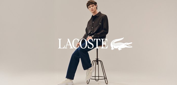 Jeune homme assis sur un tabouret, portant une chemise noire à boutons, un jean bleu foncé et des baskets blanches sur fond beige uni.