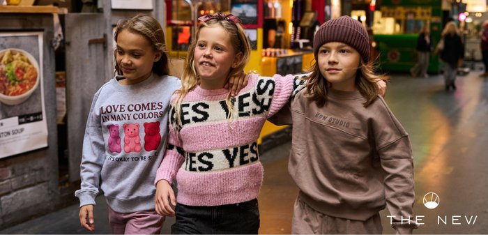 Trois enfants marchent bras dessus bras dessous dans une zone commerçante, portant des vêtements décontractés colorés et souriants.